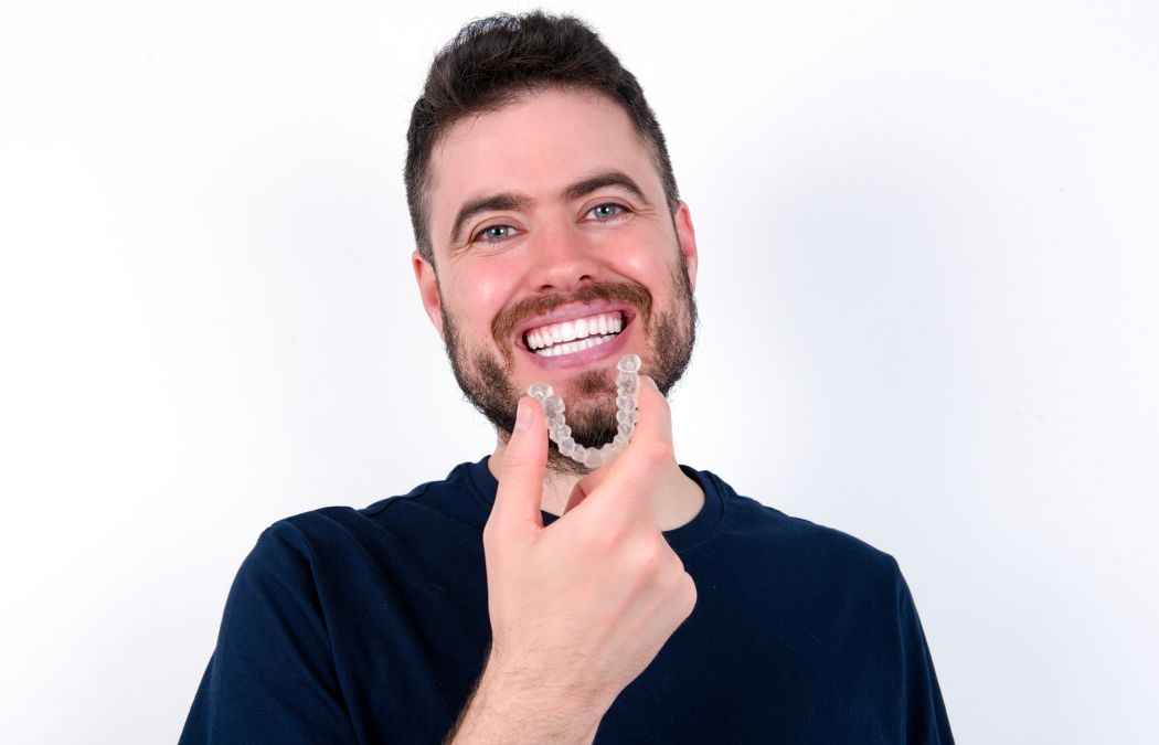 A man with a beard smiles while holding a clear dental aligner in front of his teeth against a plain white background.