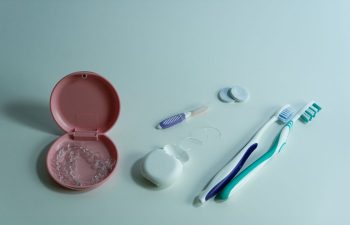 Orthodontic aligners in a pink case, a floss container, interdental brush, dental cleaning tablets, and two toothbrushes arranged on a white surface.