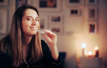 Woman with long brown hair holds a dental tray near her mouth, smiling, with lit candles and framed photos in the blurred background.