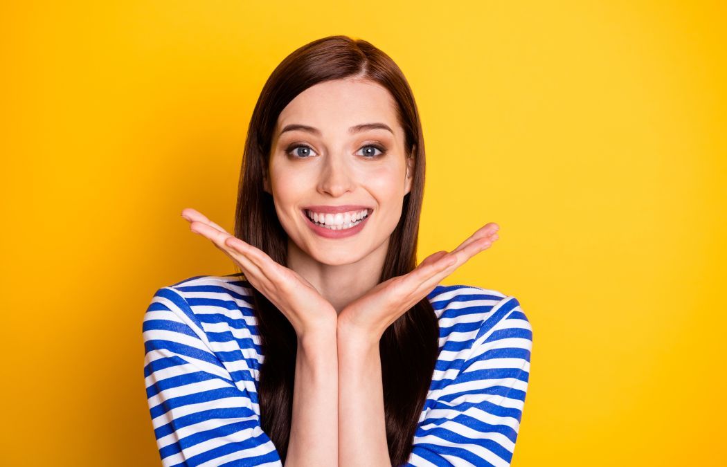 Woman with long brown hair and a striped shirt smiles, holding her hands under her chin, standing against a solid yellow background.