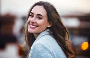 A woman with long brown hair and a light gray coat smiles while looking over her shoulder outdoors, with a blurred background.
