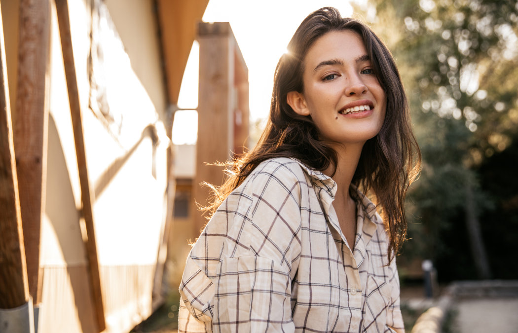 A young woman with long brown hair, wearing a plaid shirt, smiles outdoors in sunlight with trees and a building in the background.