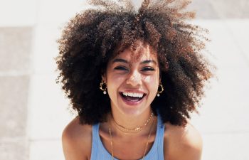 A young woman with curly hair and gold jewelry smiles broadly, wearing a light blue sleeveless top and layered necklaces, outdoors in bright sunlight.