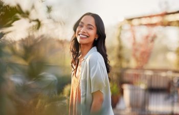 A woman with long dark hair smiles outdoors in natural light, standing among plants with a blurred background of trees and a pergola.
