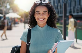 A young woman with curly hair smiles outdoors, holding notebooks and wearing a backpack. She stands on a sunny city street with people and buildings in the background.