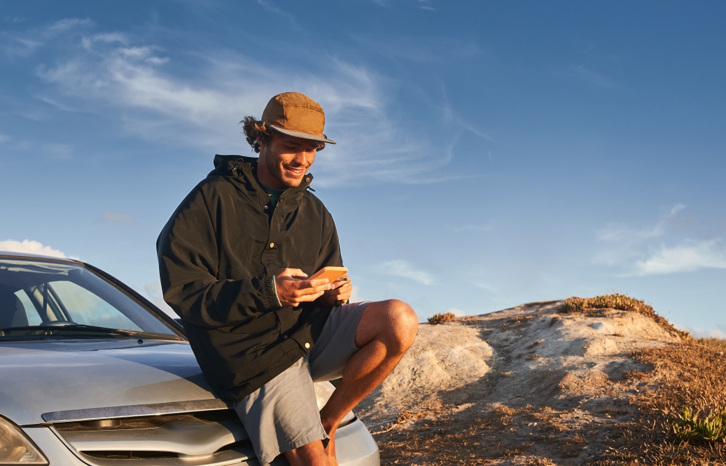 Handsome young man using mobile phone while sitting at the car at the wild beach and messaging with his friends. People and technologies concept